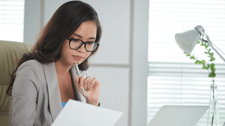 woman at her desk with laptop, pondering over a document with her pen on her chin.